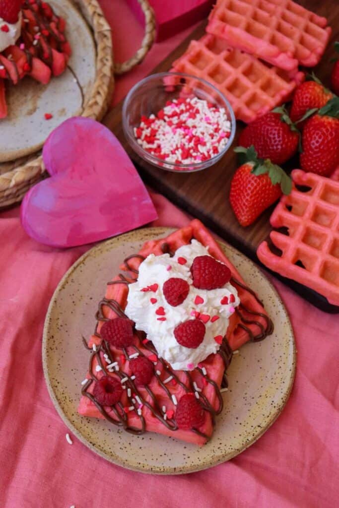 Plate of pink waffles with whipped cream, chocolate sauce, raspberries and heart sprinkles.