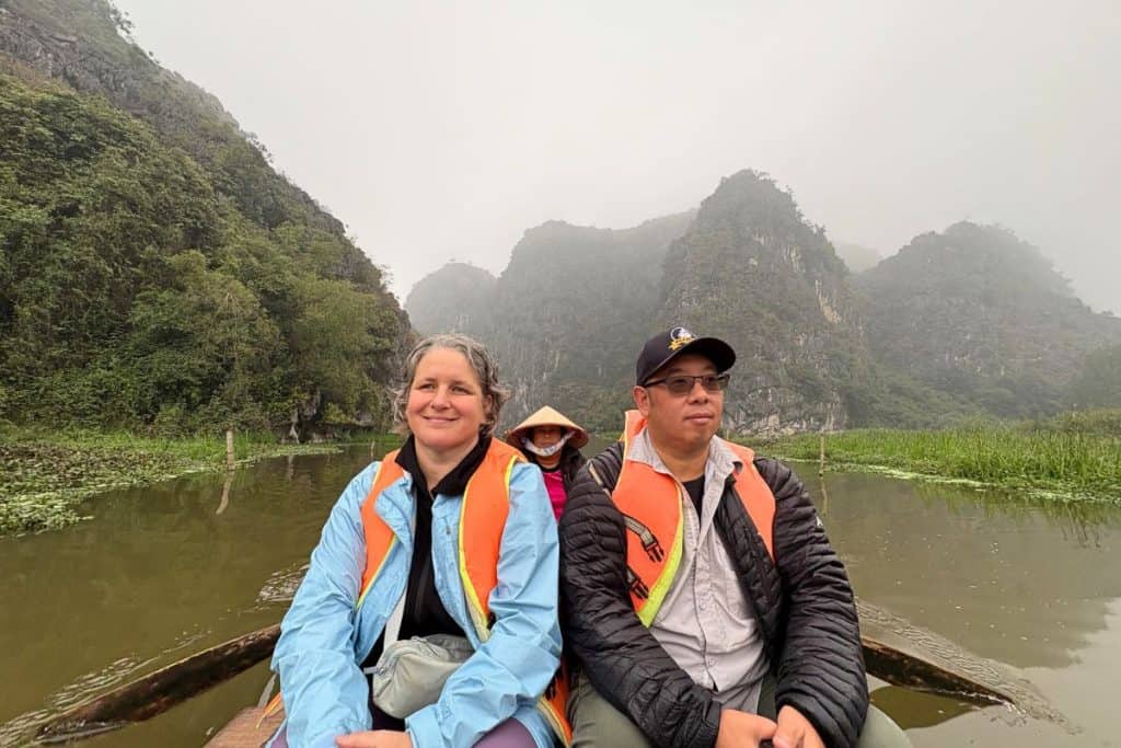 Two tourists wearing life jackets sit at the front of a wooden boat gliding through a calm river surrounded by lush greenery and misty limestone mountains. Behind them, a local rower in a conical hat and pink jacket paddles the boat, creating a peaceful and scenic moment in a rural Vietnamese landscape in the Van Long Nature Reserve.