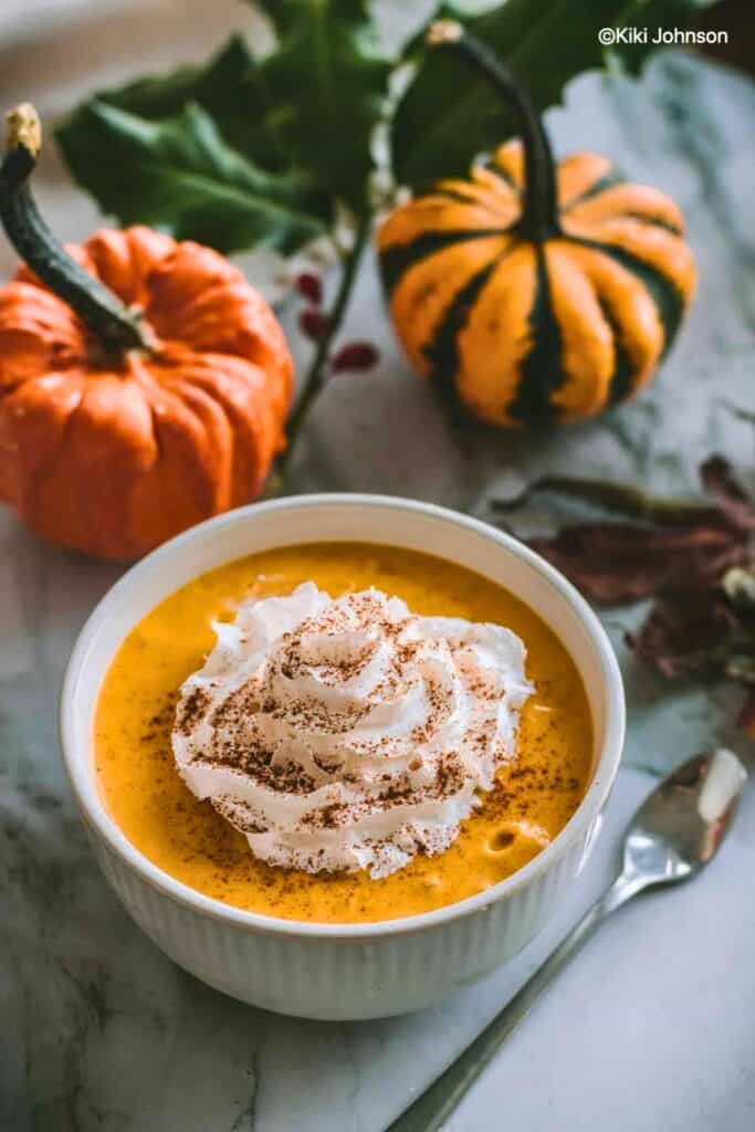 Creamy vegan pumpkin mousse topped with a swirl of whipped cream and a dusting of warm spices. Small orange pumpkins and autumn leaves create a cozy fall setting around this easy fall dessert.