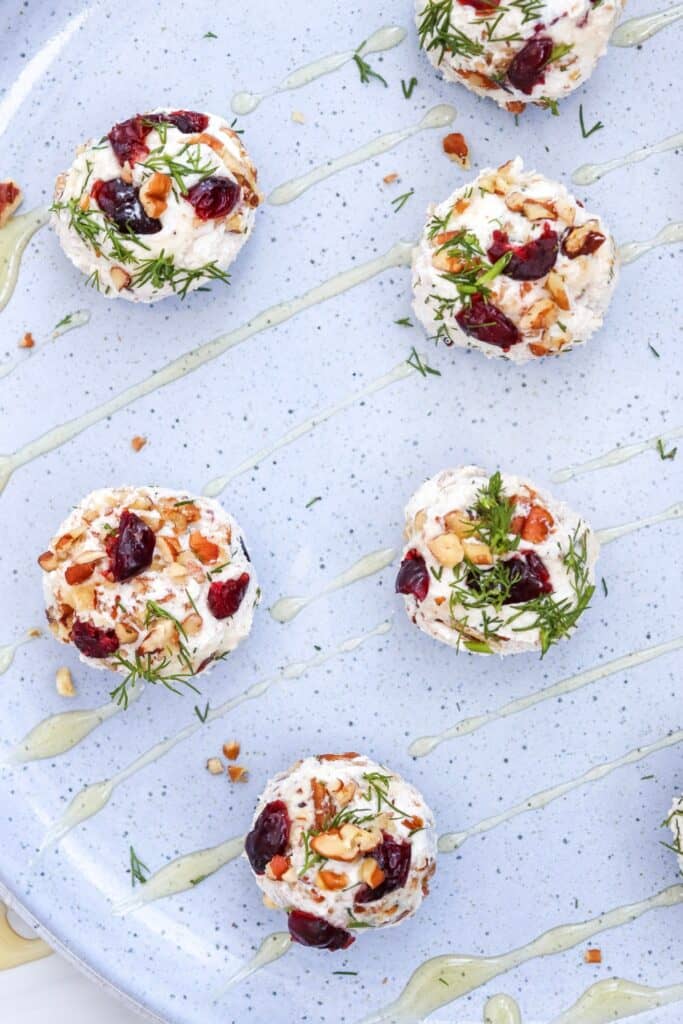 Round cranberry pecan cheese balls are spaced across a pale blue platter and topped with dark red dried cranberries. Chopped pecans and fresh green dill add color and texture to the vegetarian appetizers while thin lines of golden honey are drizzled across the platter.
