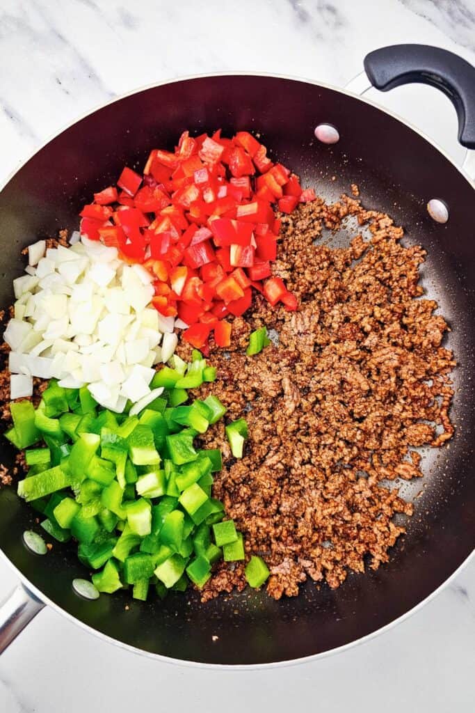 Seasoned ground beef in a skillet with freshly chopped red bell peppers, green bell peppers, and onions arranged on top, ready to be saut&eacute;ed together for sheet pan dinner quesadillas.