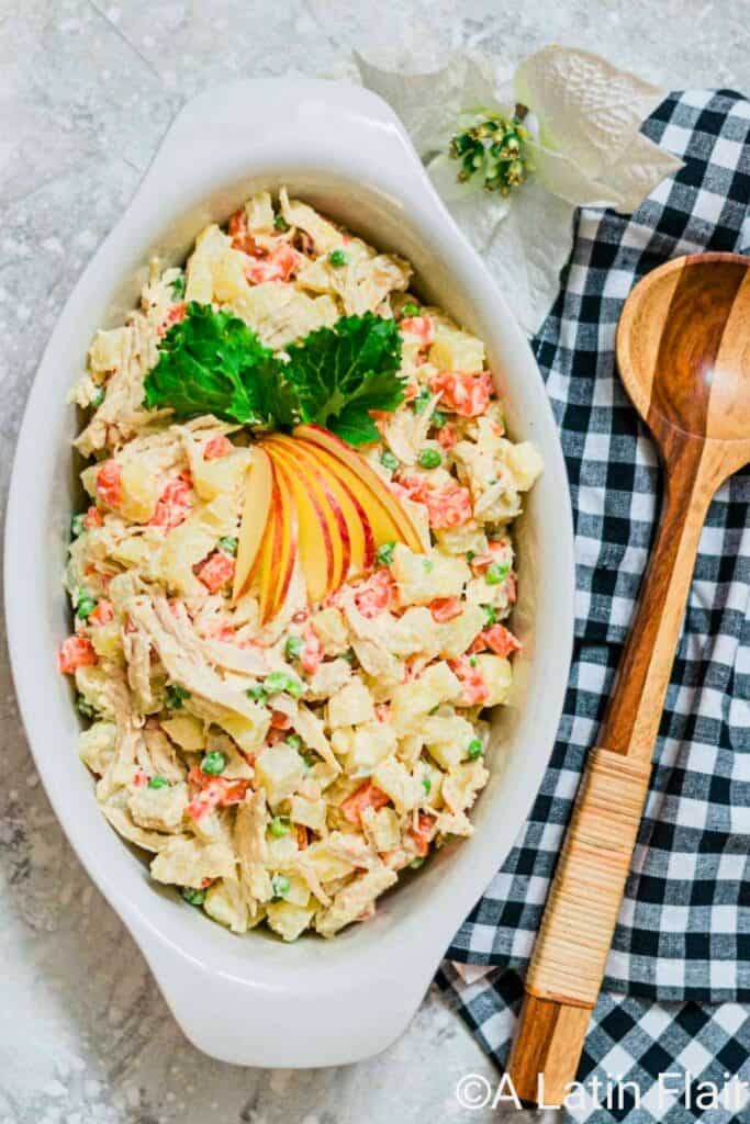 An overhead view of creamy Venezuelan Chicken Potato Salad with shredded chicken, chunks of potato, diced carrots, green peas, and a mayonnaise dressing, garnished with sliced red apple and leafy greens in a white serving dish.