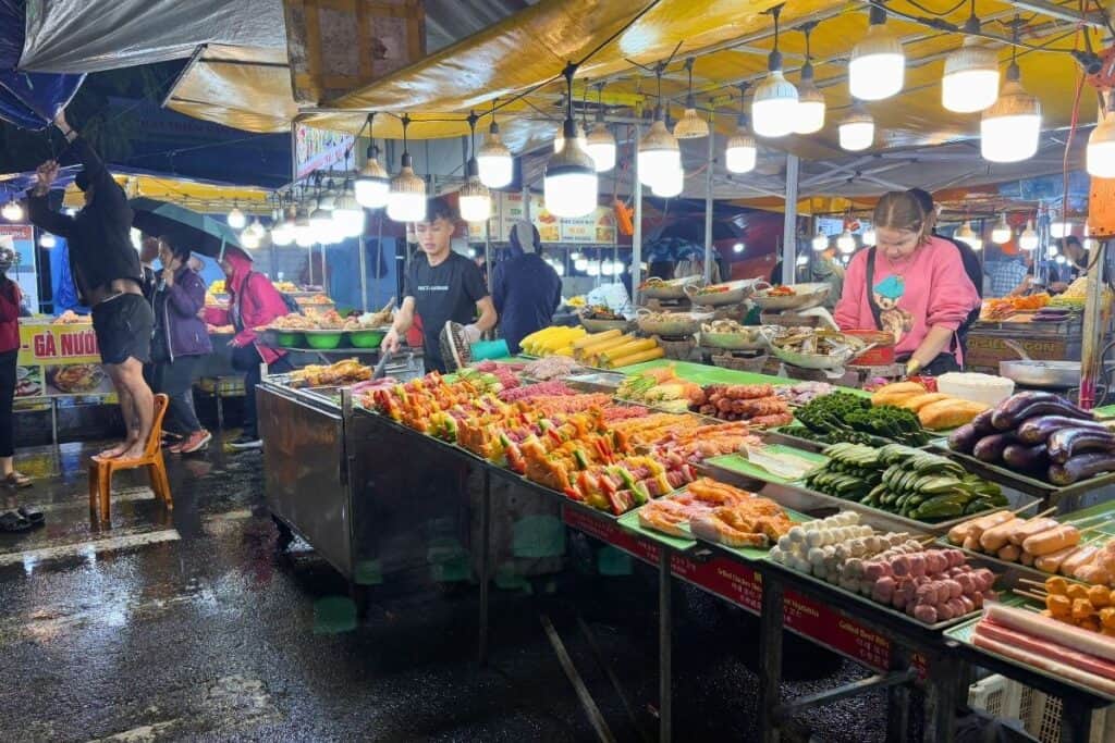 A bustling Vietnamese street food market in Da Nang, brightly lit with hanging bulbs, showcases a colorful array of skewered meats, fresh vegetables, corn, eggplant, and seafood on trays. Vendors prepare food behind the stalls while customers browse under rain-dampened tarps on a wet evening.