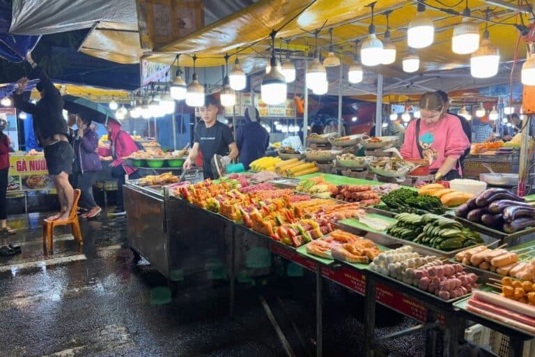 A bustling Vietnamese street food market in Da Nang, brightly lit with hanging bulbs, showcases a colorful array of skewered meats, fresh vegetables, corn, eggplant, and seafood on trays. Vendors prepare food behind the stalls while customers browse under rain-dampened tarps on a wet evening.
