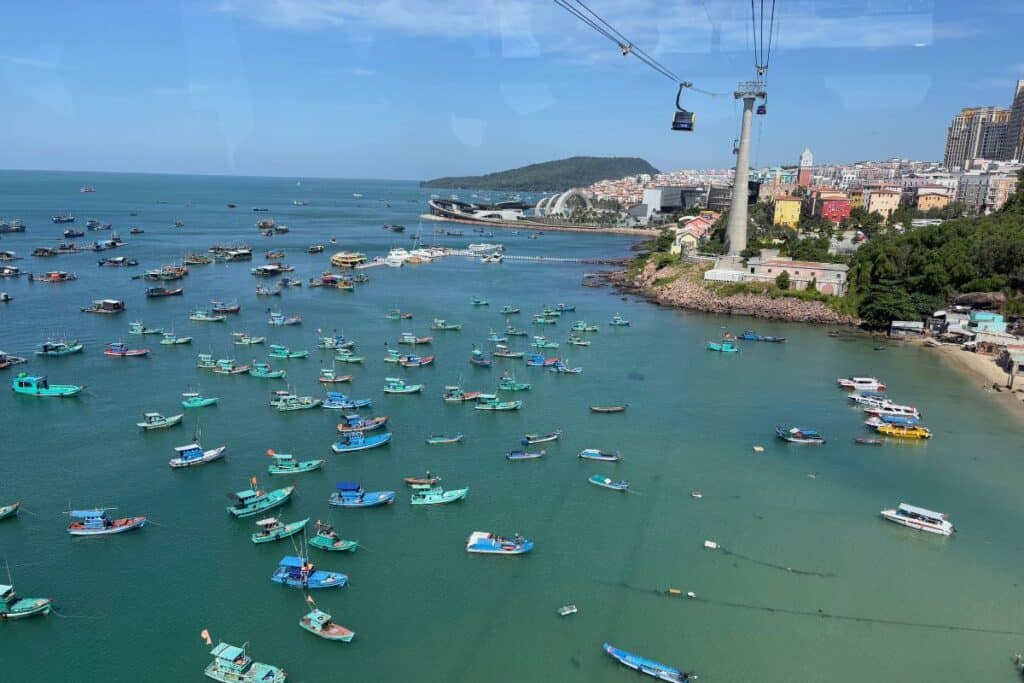 Colorful fishing boats float in turquoise waters near a bustling coastal city on Phu Quoc Island, with a cable car crossing overhead and modern buildings and lush hills in the background.