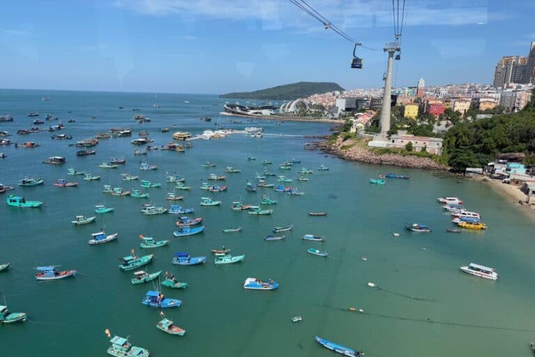 Colorful fishing boats float in turquoise waters near a bustling coastal city on Phu Quoc Island, with a cable car crossing overhead and modern buildings and lush hills in the background.