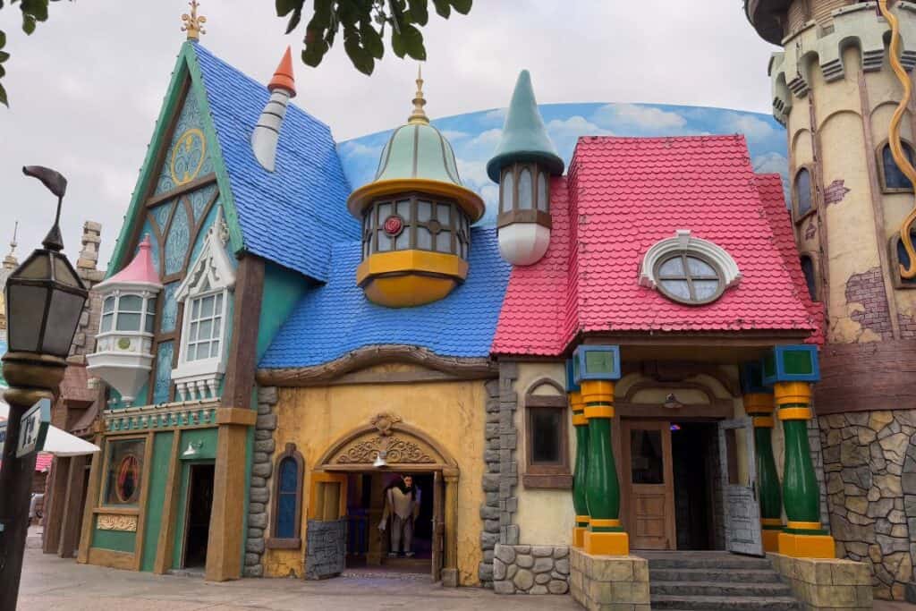 A whimsical fairytale-themed building in bright colors stands in VinWonders Fantasy Land, Phú Quốc, Vietnam. It features steep blue and red tiled roofs, turret-like windows, and ornate architectural details that evoke a playful, storybook charm. Two costumed characters can be seen inside the open yellow arched entrance.