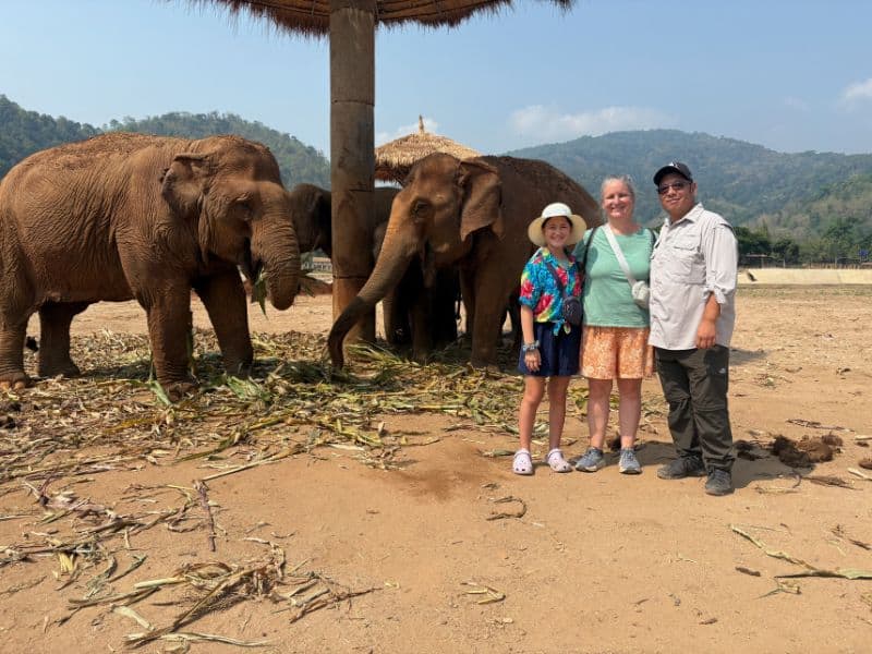 Three people pose for a photo in front of a group of elephants eating under thatched umbrellas at a wildlife sanctuary. The dry, open landscape is surrounded by forested hills under a sunny blue sky, creating a scenic and peaceful setting.