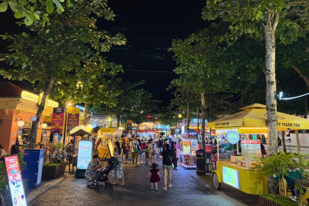 Bustling scene at the Vui Fest Night Baazar Sunset Town Phu Quoc, with families and tourists strolling under string lights and trees, surrounded by colorful food stalls and vendor carts. Bright signage and a festive atmosphere light up the night in this lively outdoor market.