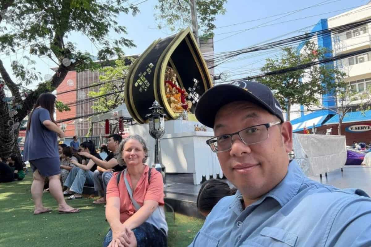 Man wearing glasses and a black cap takes a selfie while sitting on grass outside with others after an earthquake in Bangkok Thailand. Behind the group is a small shrine structure and city buildings with power lines overhead showing people gathered in an open area for safety.