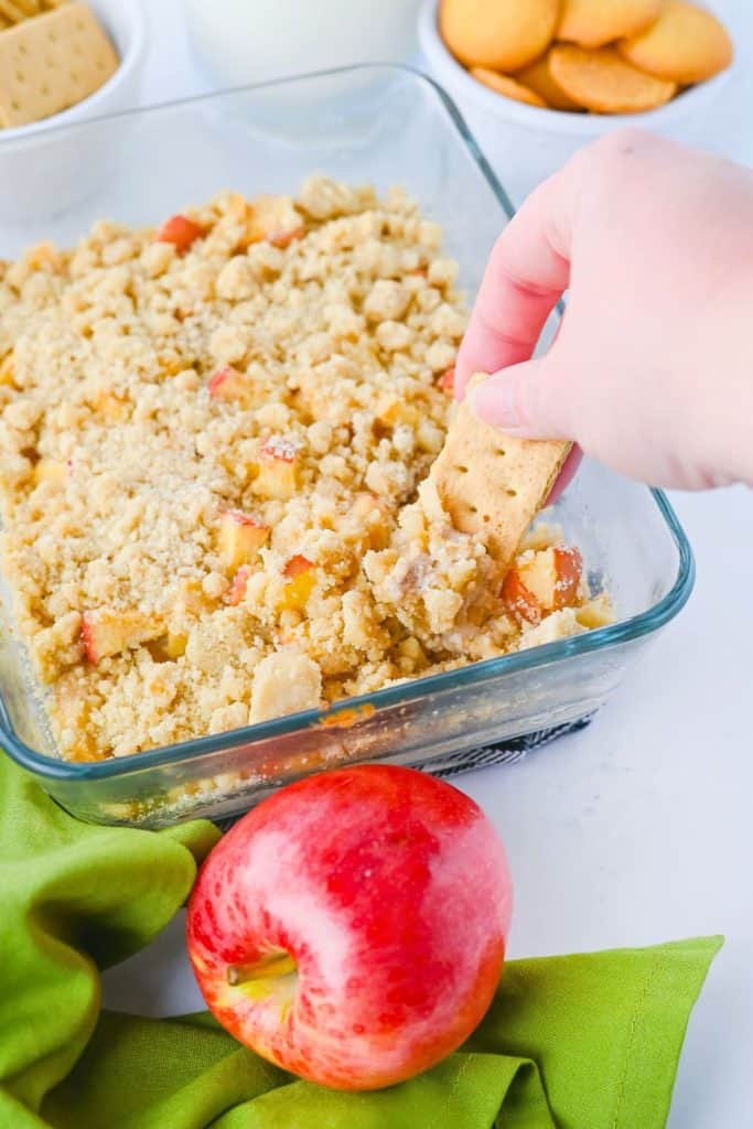 A hand dips a graham cracker into a warm apple pie dip topped with golden, crumbly streusel in a glass baking dish. A fresh red apple and green napkin sit in the foreground, with vanilla wafers and milk in the background, creating a cozy fall movie night snack vibe.