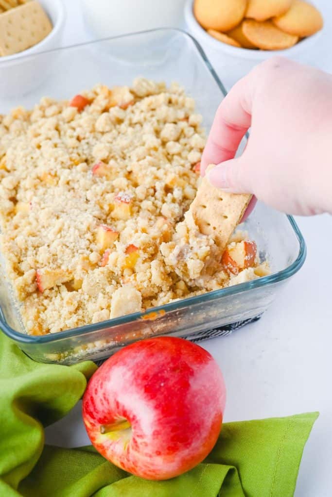 A hand dips a graham cracker into a warm, crumbly apple pie dip in a glass dish. The dip is surrounded by a fresh red apple, green cloth, and bowls of cookies and crackers, making it a cozy fall appetizer or dessert dip.