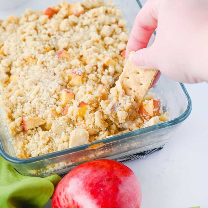 A hand dips a graham cracker into a warm, crumbly apple pie dip in a glass dish. The dip is surrounded by a fresh red apple, green cloth, and bowls of cookies and crackers, making it a cozy fall appetizer or dessert dip.