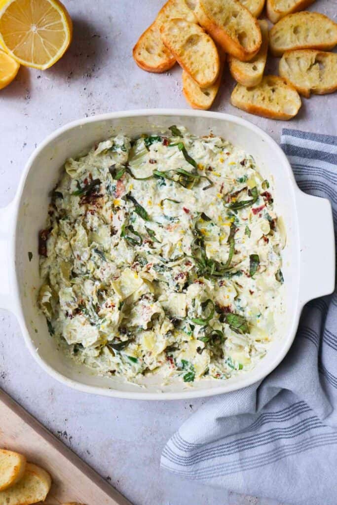 Warm Boursin cheese dip with artichoke pieces, sun dried tomatoes, and fresh herbs is served in a white baking dish. Toasted baguette slices and fresh lemon sit nearby for serving with the creamy dip.