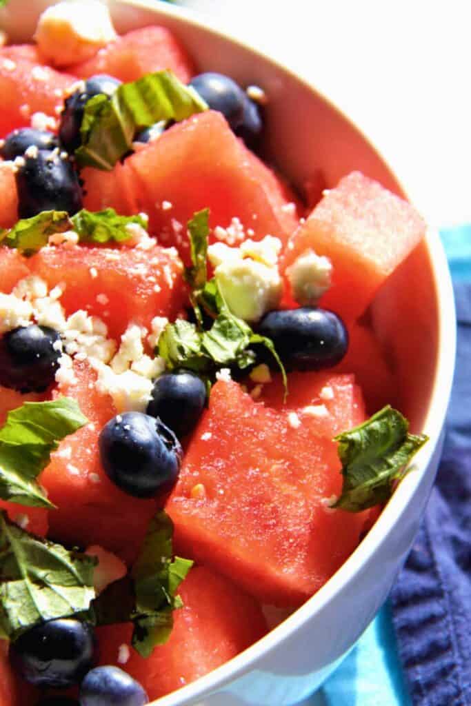Overhead view of half a low bowl of watermelon, feta, blue berry salad on top of a blue dish towel. 