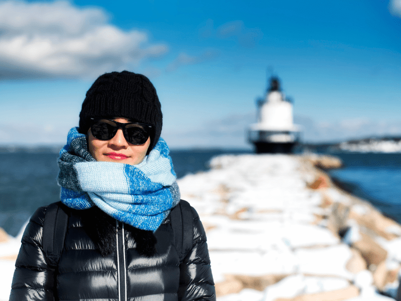 What to wear in Maine in winter. Chinese woman wearing down jacket, hat and scarf in front of ocean and Spring Point Ledge lighthouse. 