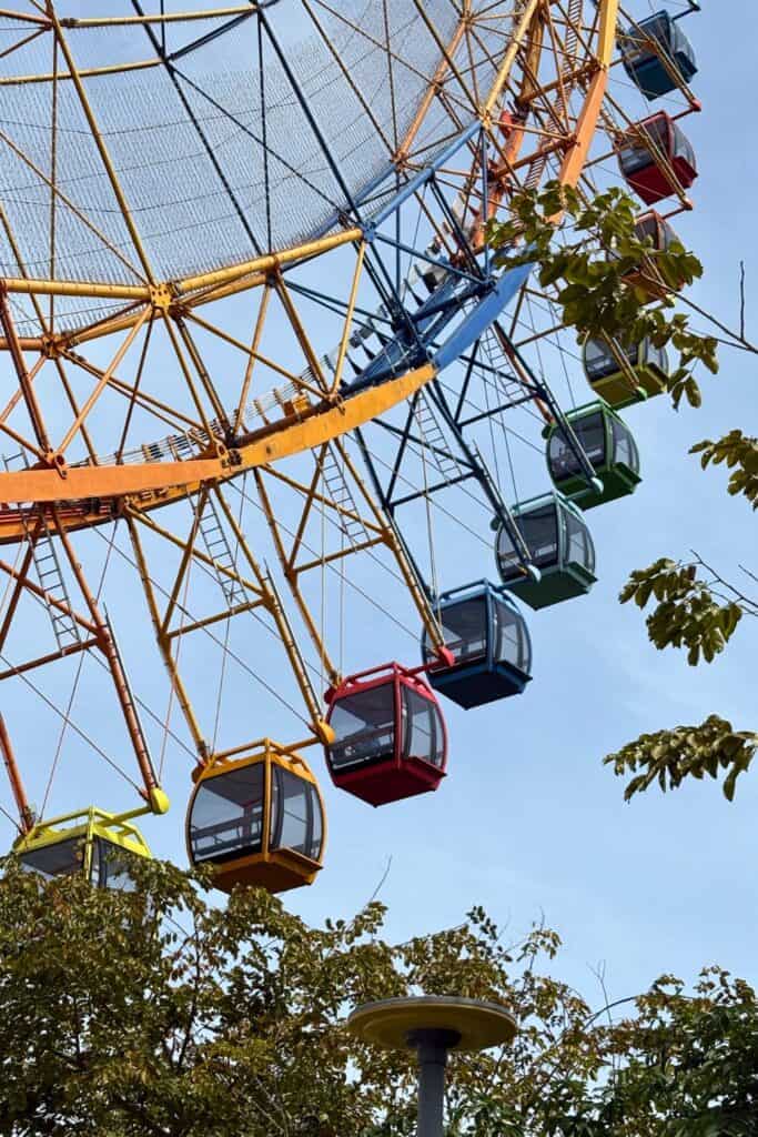 Close-up of the colorful Wheel of Time Ferris wheel at VinWonders Phu Quoc, with bright red, yellow, green, and blue gondolas rising above treetops against a clear blue sky.