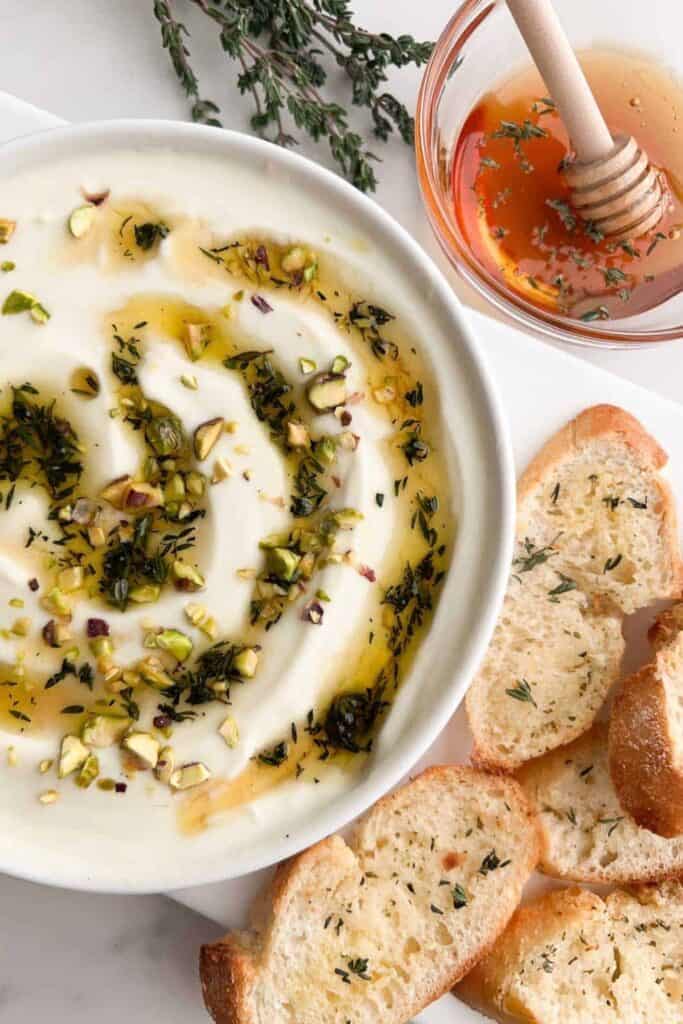 Creamy whipped ricotta dip drizzled with honey and topped with chopped pistachios and fresh thyme. Toasted bread slices and a small bowl of honey are served alongside the dip.