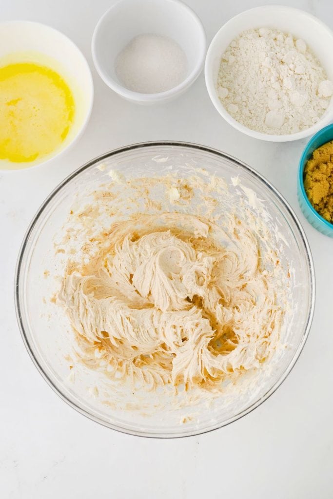 A glass mixing bowl filled with whipped cream cheese mixture, lightly swirled and spiced for apple cream cheese dip. Surrounding it are small bowls of melted butter, white sugar, flour, and brown sugar, laid out on a white surface.