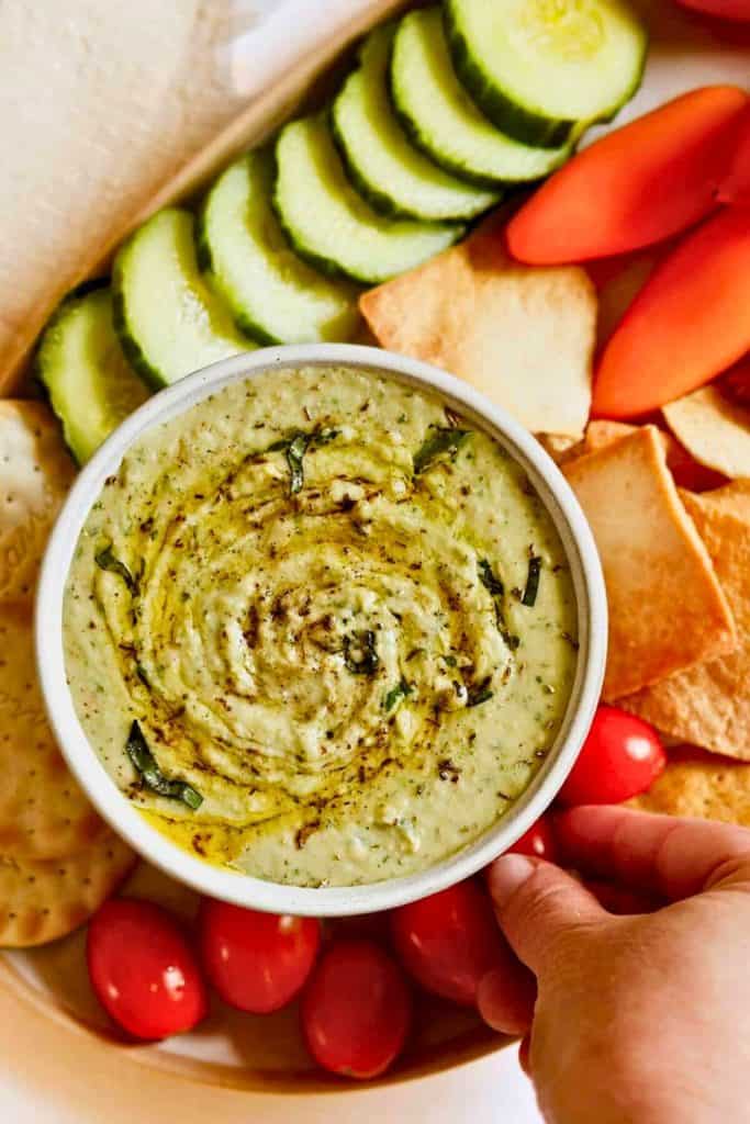 Creamy White Bean Basil Dip in a white bowl, swirled with olive oil and sprinkled with fresh basil and black pepper. Served on a platter with cucumber slices, grape tomatoes, pita chips, and crackers for dipping.