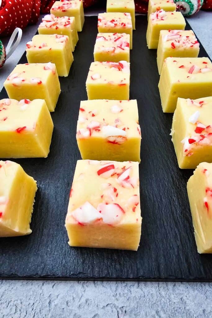Rows of creamy yellow, white chocolate fudge squares topped with crushed candy canes are neatly arranged on a black slate tray. This easy homemade Christmas fudge is festive and inviting, with red, green  and white holiday decor in the background.