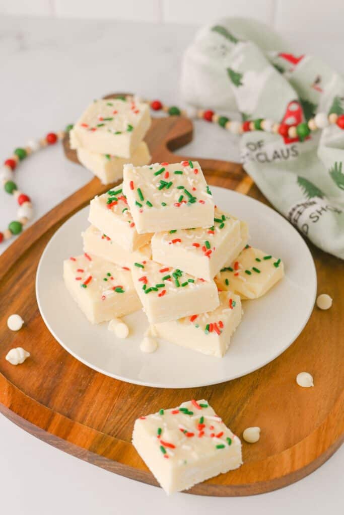 A plate stacked with squares of White Chocolate Christmas Fudge topped with red, green, and white sprinkles, surrounded by white chocolate chips and festive holiday decorations.