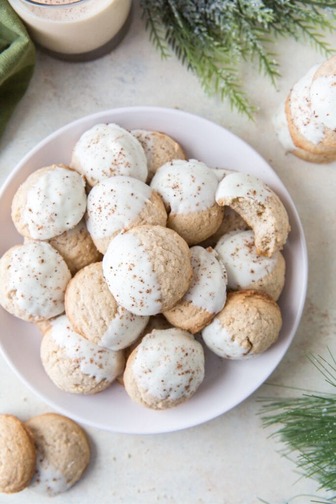 Plate of soft, round eggnog cookies partially dipped in white chocolate and sprinkled with ground nutmeg. The cookies have a festive holiday feel, surrounded by evergreen branches and a glass of eggnog in the background.