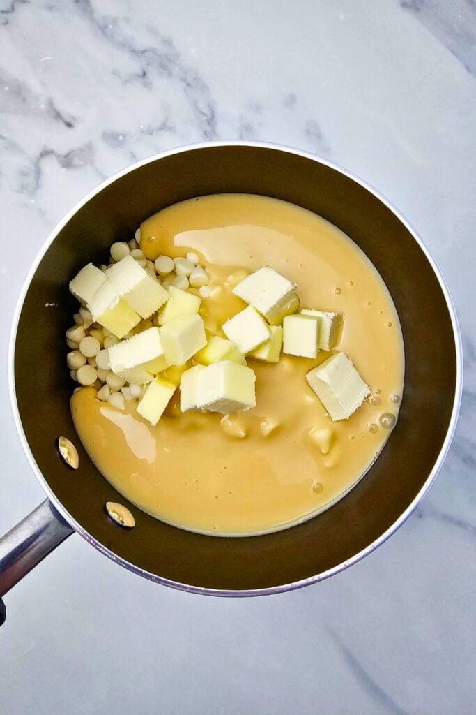 White chocolate chips, sweetened condensed milk, and cubed butter are combined in a saucepan on a marble countertop. These rich ingredients are the first step in making creamy homemade peppermint fudge.