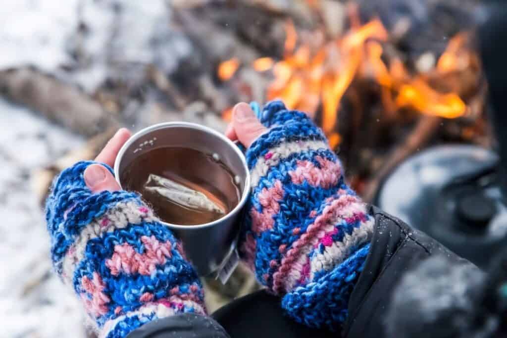 knitted fingerless mitts holding cup of tea, with lit fire in background.