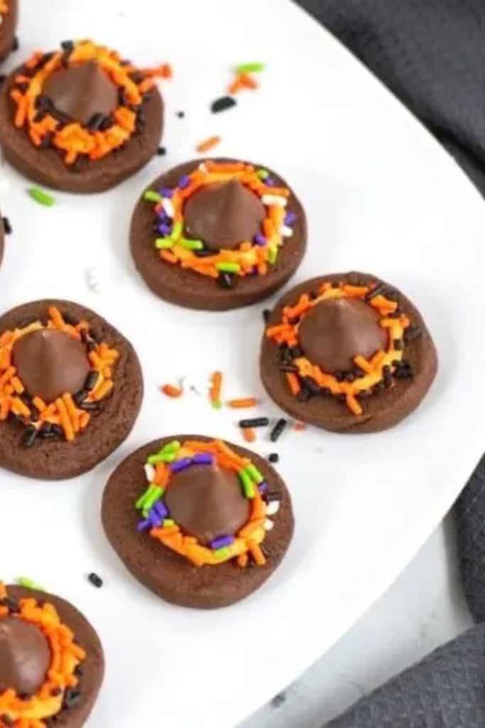 Chocolate thumbprint cookies topped with a Hershey's Kiss and decorated with Halloween-colored sprinkles in orange, purple, green, black, and white. Arranged on a white plate for a festive and colorful Halloween treat.