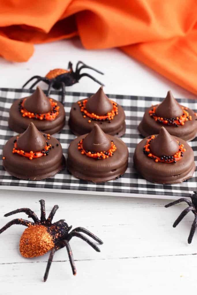 Chocolate witch hat cookies made from chocolate-covered sandwich cookies topped with a chocolate kiss, decorated with orange icing and black-and-orange sprinkles. Arranged on a checkered tray with glittery Halloween spider decorations and an orange fabric background.