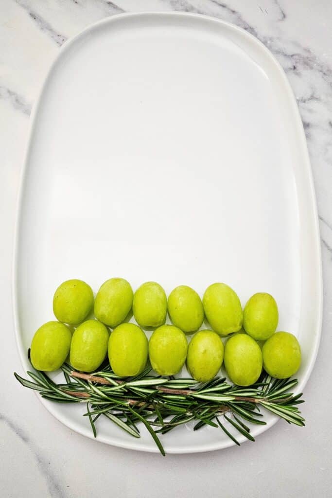 Two neat rows of green grapes arranged above fresh rosemary sprigs on a white oval serving tray, showing the first step in creating easy Xmas fruit platter ideas for holiday potlucks.