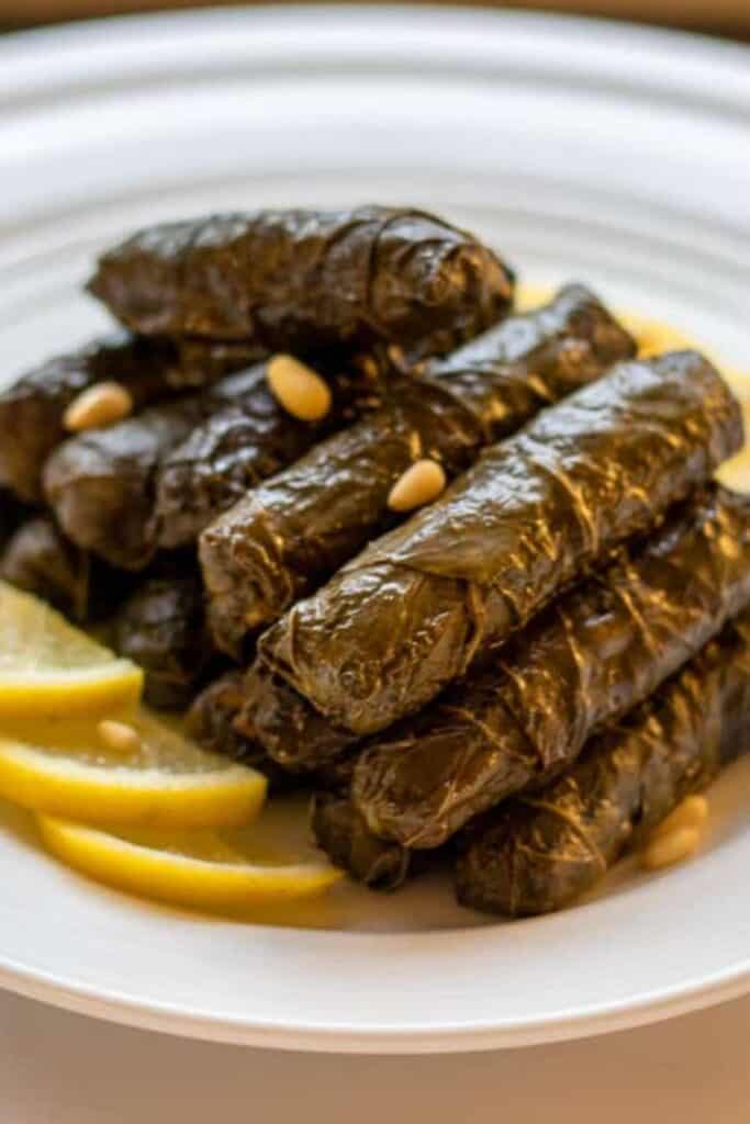 Plate piled with rolls of stuffed grape leaves, with sliced lemon on the side. 