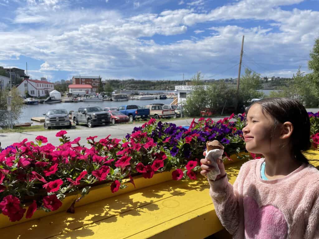 young girl eating an ice cream cone in front of colorful flowers, with a lake and blue sky with clouds in background. Yellowknife, NT, Canada