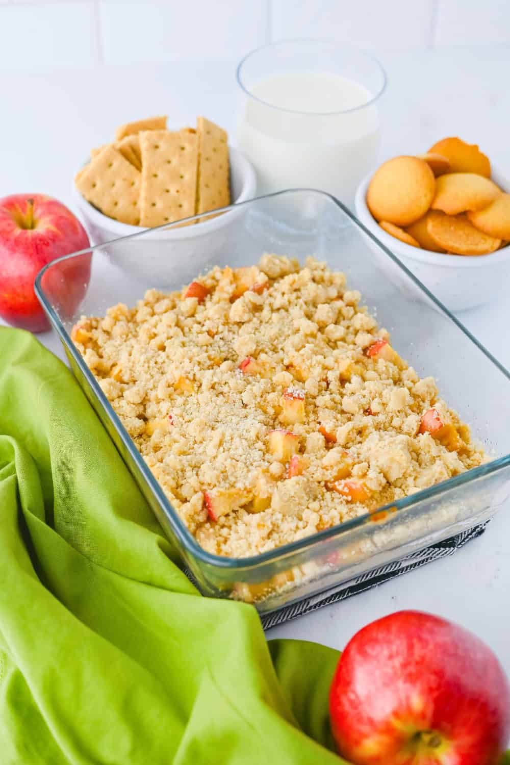 Apple pie dip made with chunks of red apple and topped with a golden crumbly streusel in a glass baking dish. Graham crackers and vanilla wafers are served alongside the dip with fresh red apples and a glass of milk.