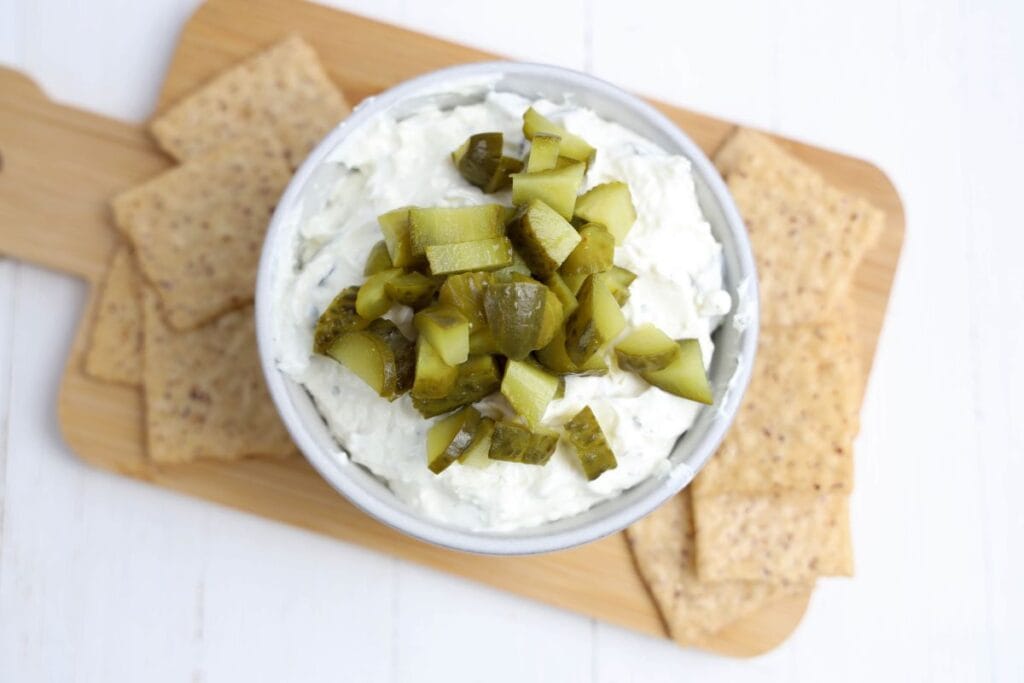 Overhead view of a bowl of thick and creamy dill pickle dip topped with chopped pickles, served on a wooden board surrounded by rectangular whole grain crackers. 