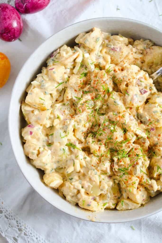 Creamy potato salad in a white bowl, made with chunks of potato, diced pickles, red onion, and hard-boiled egg, all mixed in a seasoned mayo-based dressing. The salad is garnished with fresh herbs and a sprinkle of paprika for color.