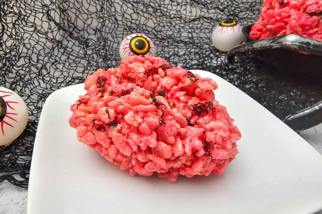 Close-up of a pink rice krispie treat shaped like a brain with dark vein-like details, displayed on a white plate with spooky eyeball decorations and a black net backdrop.