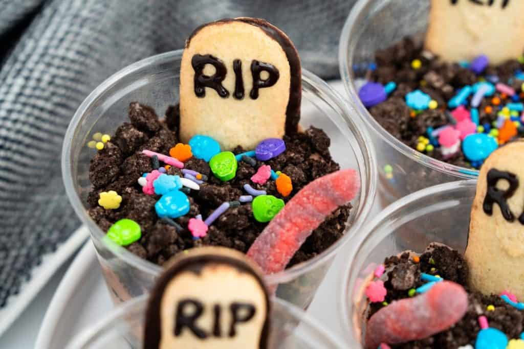 Plastic cups filled with chocolate pudding and crushed cookies to mimic dirt, each decorated with a cookie tombstone marked “RIP,” bright Halloween sprinkles, and gummy worms for a zombie-themed dessert. The setup creates a playful, graveyard-style look perfect for Halloween parties.