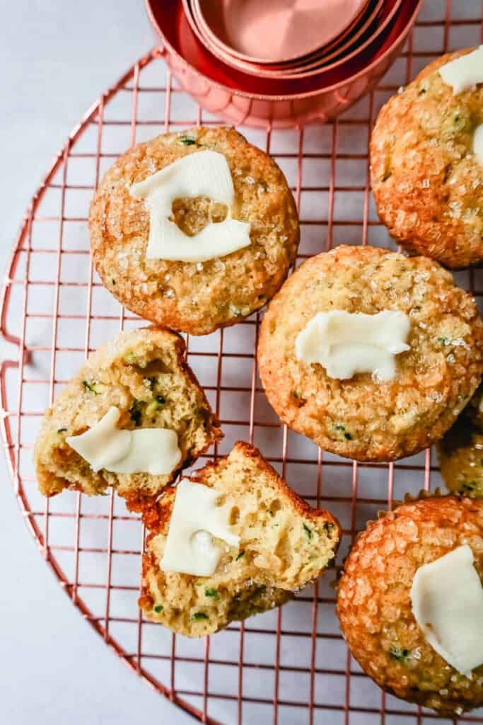 Golden zucchini muffins topped with melting pats of butter and a sprinkle of coarse sugar, cooling on a copper wire rack. One muffin is split open to reveal flecks of green zucchini in the moist interior, making them a wholesome and portable snack perfect for camping.