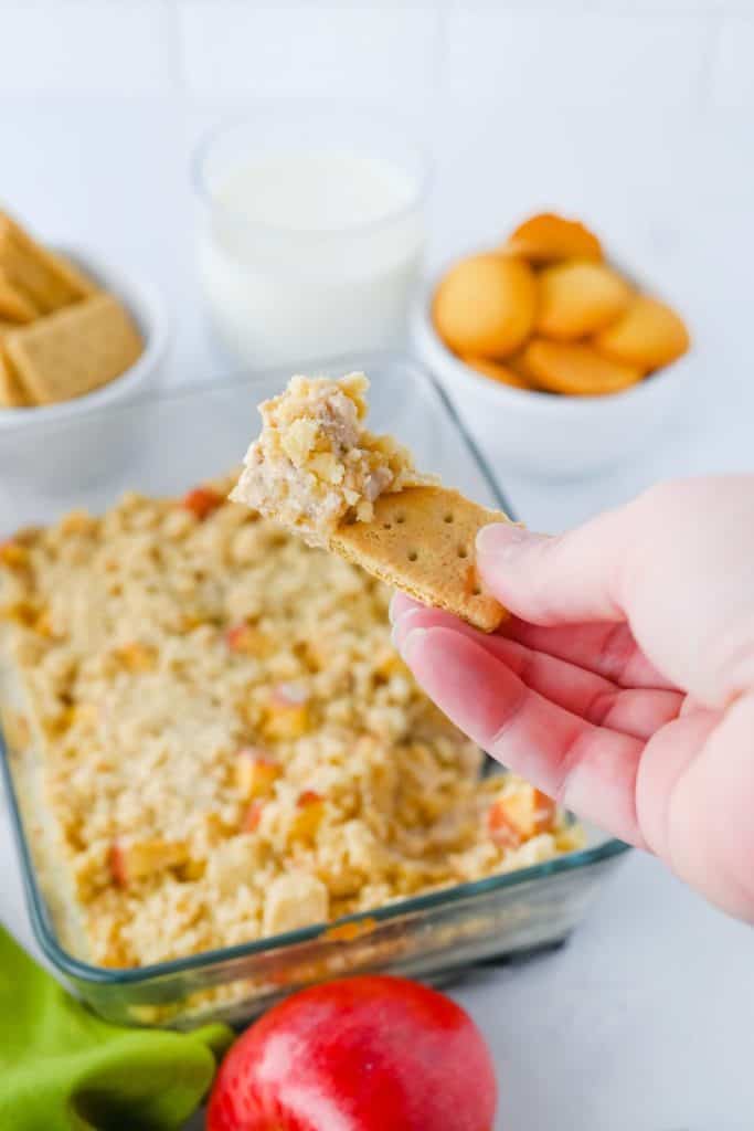 A hand holds a graham cracker topped with a scoop of cream cheese apple dip, showing creamy filling and crumbly topping. In the background, the dip sits in a glass dish alongside a red apple, vanilla wafers, graham crackers, and a glass of milk.