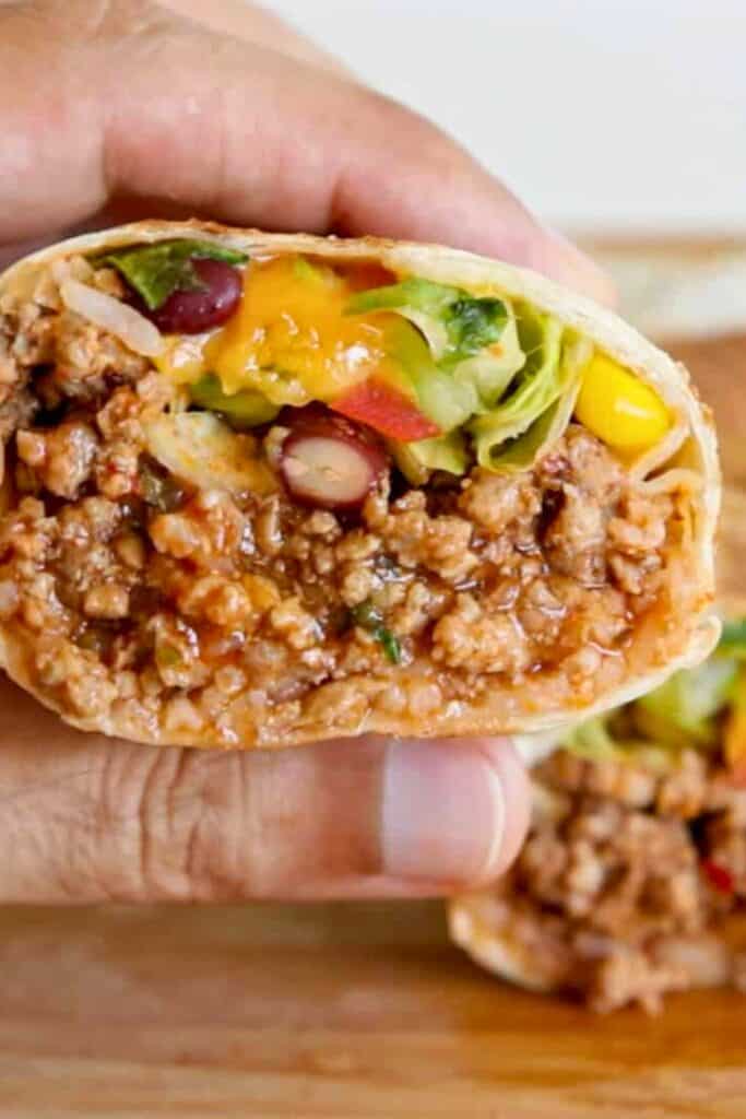 Beef burrito held in hand showing a soft tortilla filled with seasoned ground beef, rice, beans, corn, melted cheese, and lettuce. Close up of the burrito cross section with the saucy beef filling and colorful toppings packed inside.