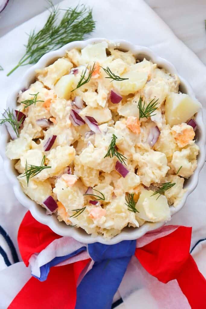 Close up overhead view of a scalloped bowl filled with classic potato salad with egg and may dressing, decorated with a red white and blue bow. 
