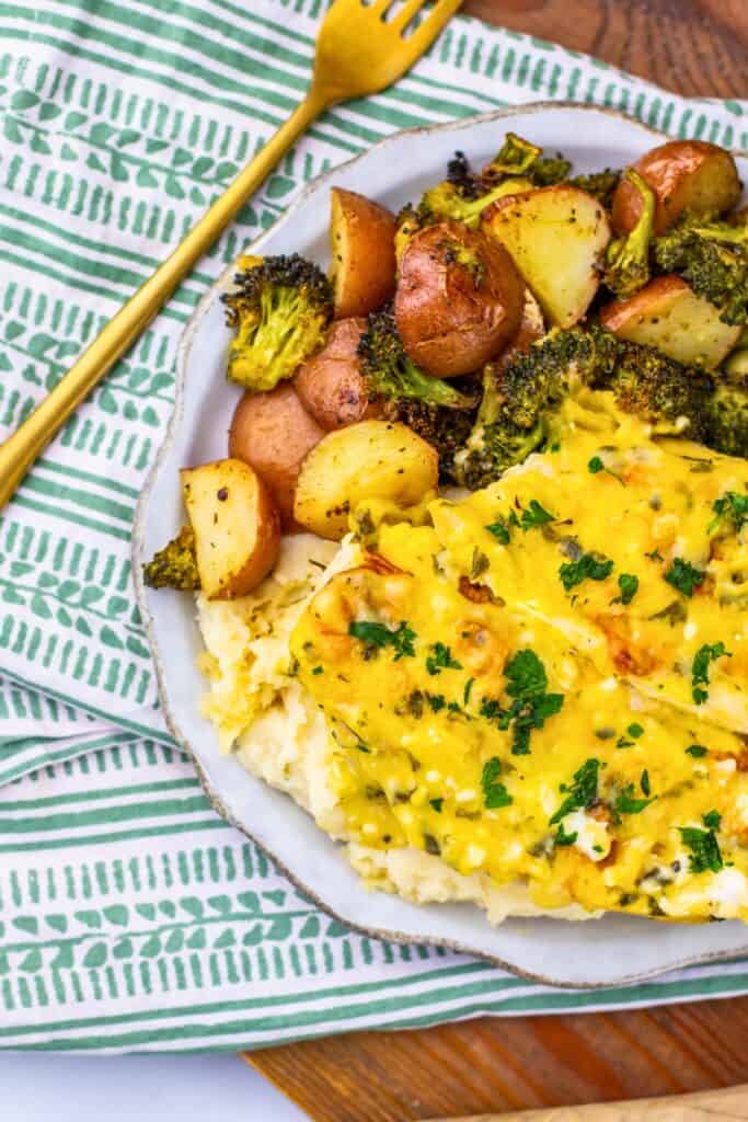 Overhead view of half a plate of chicken and potatoes sheet pan dinner served over mashed potatoes, topped with creamy parmesan sauce and chopped parsley, alongside roasted red potatoes and broccoli. The dish is plated on a ceramic plate over a green and white striped napkin.