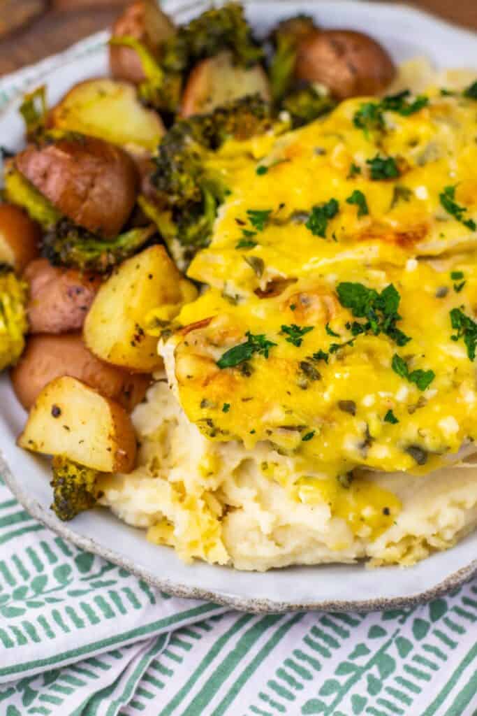 Close-up of a chicken breast sheet pan dinner with cheesy garlic parmesan chicken over mashed potatoes, served with roasted red potatoes and crispy broccoli. The food is plated on a rustic dish over a green and white patterned napkin.