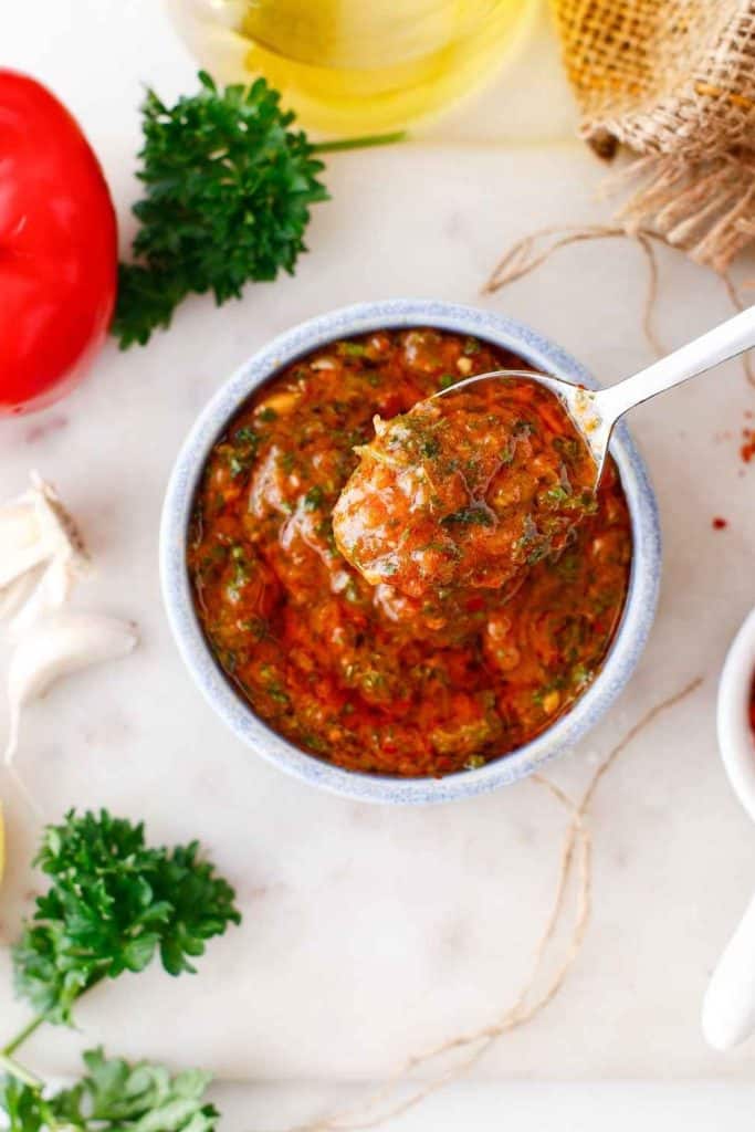Vibrant Chimichurri Rojo served in a small bowl, with a spoon lifting the chunky red and green herb sauce. Surrounded by fresh parsley, garlic, and a red pepper, this unique tailgate party dip bursts with bold flavor and color.