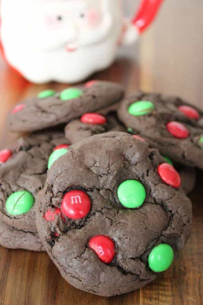 Thick, fudgy chocolate cake mix cookies studded with red and green holiday M&M candies. The cookies are piled on a wooden surface with a festive Santa mug blurred in the background, perfect for an easy Christmas treat.