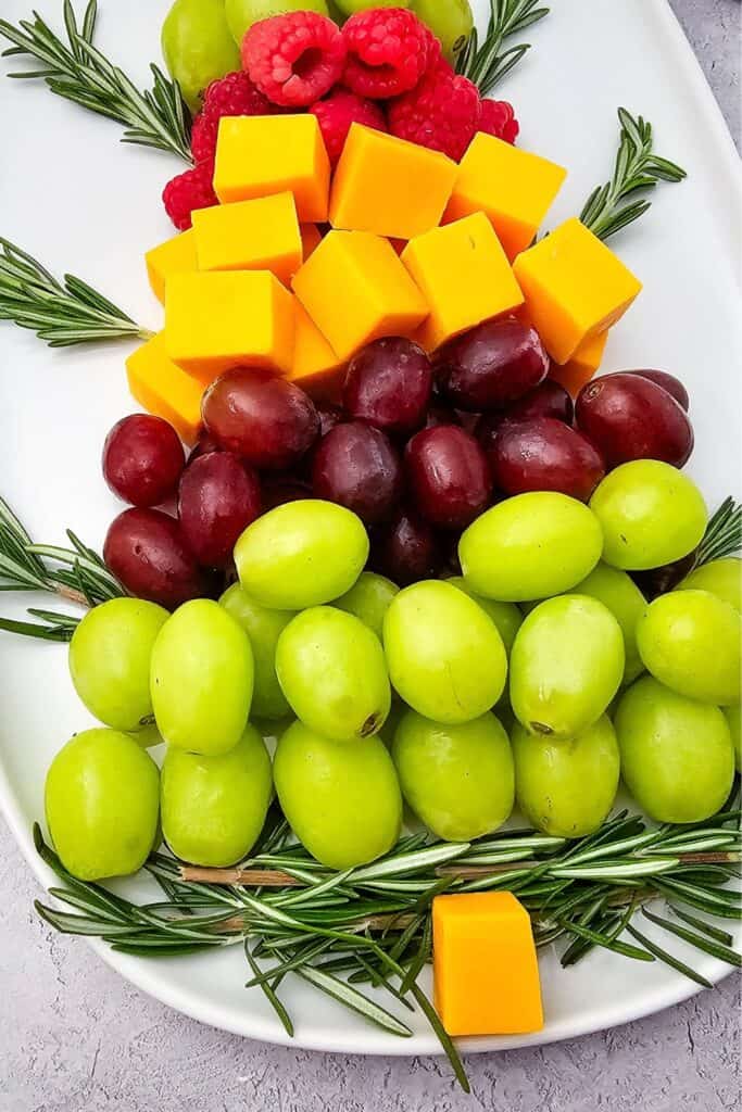 Close up of a Christmas tree charcuterie board with fruit, featuring layers of green and red grapes, fresh raspberries, cheddar cheese cubes, and rosemary sprigs arranged on a white platter with a cheese cube forming the tree trunk.