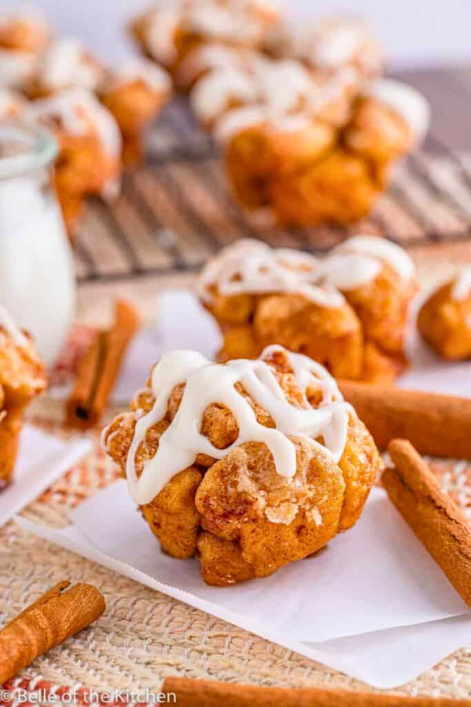 Cinnamon roll monkey bread muffins coated in cinnamon sugar and drizzled with white icing served on parchment paper with cinnamon sticks nearby. One muffin is in focus showing its pull apart texture while others cool in the background.