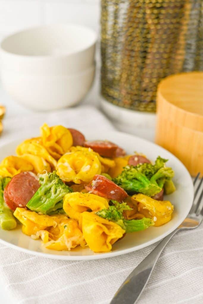 Close-up of a finished easy sheet pan meal served on a white plate, featuring cheesy tortellini, sliced smoked sausage, and roasted broccoli. The dish is colorful and hearty, resting on a soft striped napkin with a fork beside it.